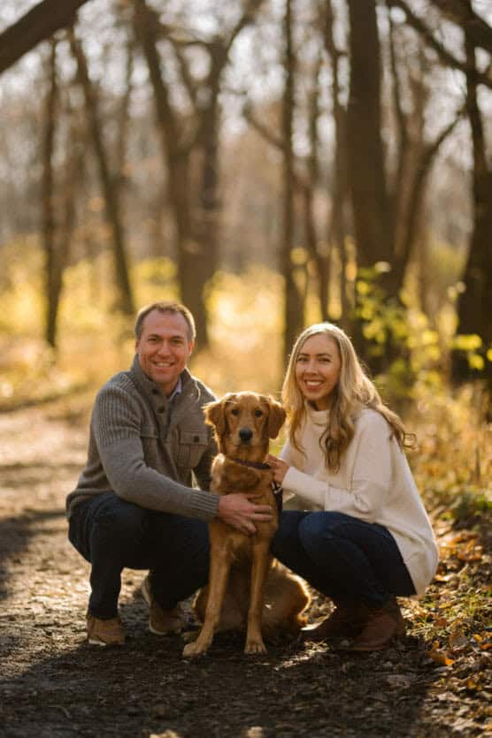 A man and a woman kneel on a sunlit forest path, smiling at the camera with a brown dog sitting between them. The autumn trees and fallen leaves create a warm, golden background. by Justin Salem Meyer