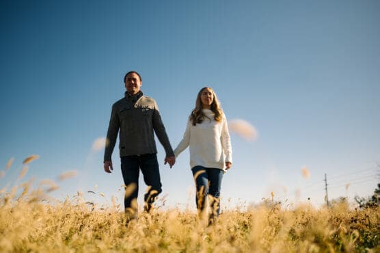 A man and woman hold hands while walking through a grassy field under a clear blue sky, with tall dry grass in the foreground and sunlight illuminating the scene. by Justin Salem Meyer