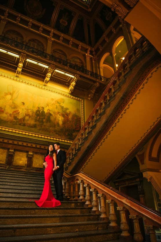 A couple stands on an ornate, grand staircase in an elegant building. The woman wears a red dress, while the man is in a dark suit. Behind them is a large mural and decorative architecture with warm lighting. by Justin Salem Meyer