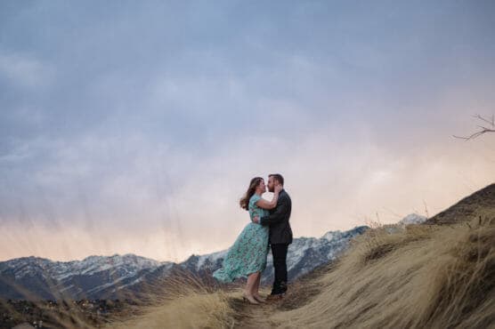 A couple stands on a grassy hillside, embracing and smiling at each other, with snow-capped mountains and a colorful sunset sky in the background. by Justin Salem Meyer