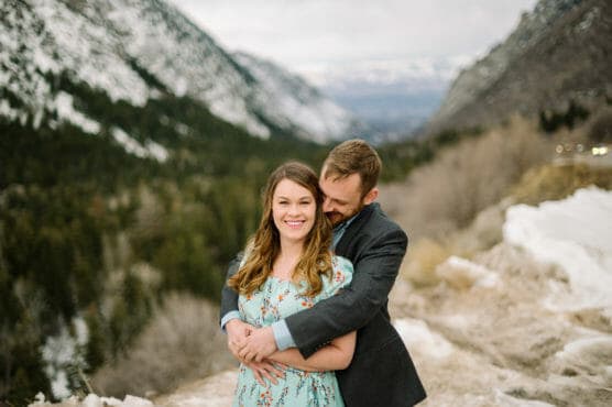 A couple stands on a snowy mountain path, with the man hugging the woman from behind. The woman smiles at the camera, while the man looks at her. Snow-covered mountains and pine trees are visible in the background. by Justin Salem Meyer