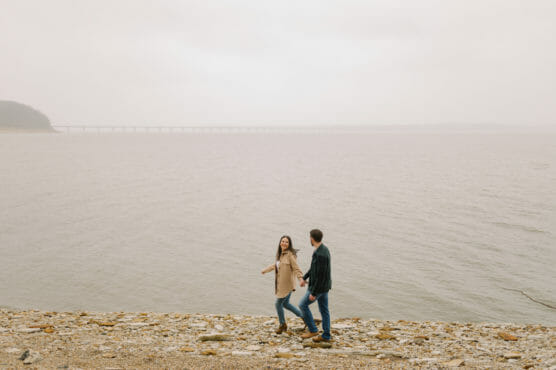 A couple walks hand-in-hand along a rocky lakeshore under an overcast sky, with a distant bridge and misty landscape in the background. by Justin Salem Meyer