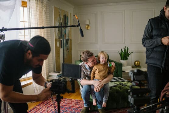 A man sits on a green ottoman in a living room, holding a young child. A camera crew with lights and microphones films them, capturing a behind-the-scenes scene on a cozy set. by Justin Salem Meyer