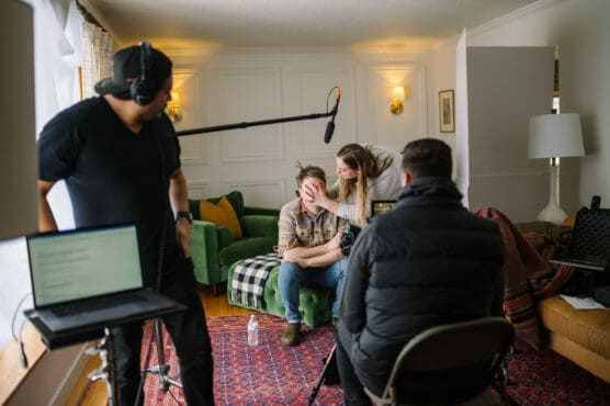 A woman applies makeup to a seated man while two crew members, one holding a boom mic, prepare for filming in a cozy living room with cameras and equipment. by Justin Salem Meyer