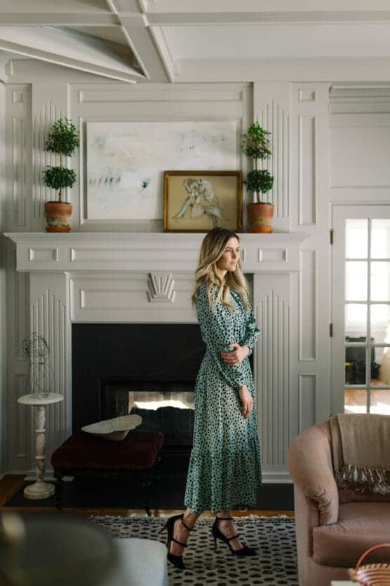 A woman in a long green polka-dot dress stands in a stylish living room by a white mantel with plants and framed art, featuring panelled walls, a fireplace, and elegant decor. by Justin Salem Meyer