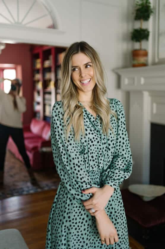 A woman with long blonde hair wearing a green dress with black polka dots stands smiling indoors, while another person in the background takes a photo of her in a living room setting. by Justin Salem Meyer