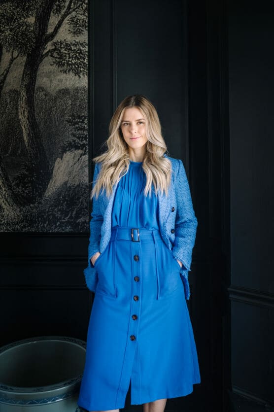 A woman with long blonde hair stands indoors, wearing a bright blue belted dress and matching textured jacket. Her hands are in her pockets, and she is standing in front of a dark wall with a black and white tree artwork. by Justin Salem Meyer