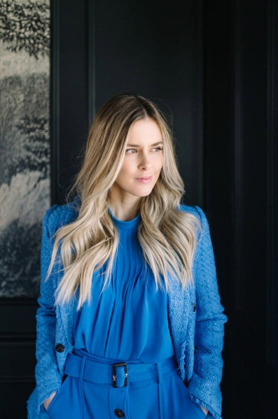 A woman with long, wavy blonde hair stands in front of a dark wall, wearing a bright blue blouse, matching jacket, and pants, looking slightly to the side with a relaxed expression. by Justin Salem Meyer