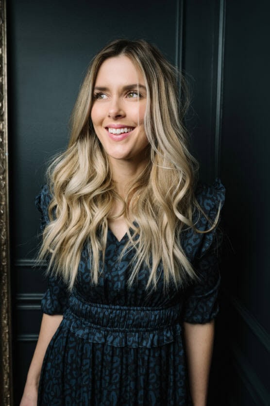 A woman with long, wavy blonde hair is smiling and looking slightly to the side. She is wearing a dark, patterned dress with puffed sleeves, standing against a dark-colored wall. by Justin Salem Meyer
