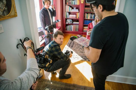 A film crew works indoors; one person holds a camera, another reviews a storyboard, while others prepare equipment. Bookshelves fill the background, and sunlight streams onto the wooden floor. by Justin Salem Meyer