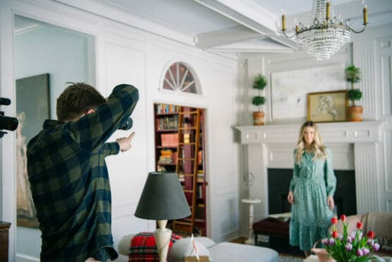 A photographer takes a picture of a woman in a green dress standing in a stylish living room with a chandelier, fireplace, bookshelves, and a vase of tulips on the table. by Justin Salem Meyer