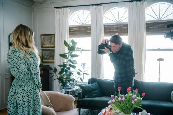 A photographer takes a picture of a woman with long blonde hair in a polka dot dress, standing in a bright living room with large windows, a green sofa, plants, and a vase of tulips on the table. by Justin Salem Meyer