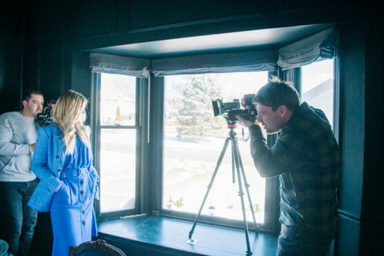 A woman in a blue outfit poses in front of a large window while two men photograph her, one using a camera on a tripod and the other holding a video camera. Snowy scenery is visible outside the window. by Justin Salem Meyer
