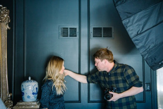 A man holding a camera leans toward a woman with long blonde hair standing against a dark blue wall, engaging her in conversation. A decorative vase and ornate mirror are visible on the left, and studio lighting is on the right. by Justin Salem Meyer