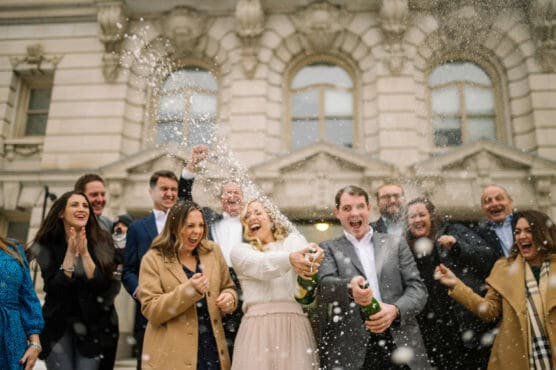 A joyful group of people stands outside a historic building, laughing as two people in the center spray champagne, with bubbly foam flying in the air. Everyone appears excited and celebratory, dressed in winter coats. by Justin Salem Meyer