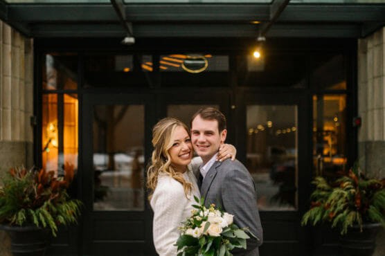 A smiling couple embraces outside a building entrance. The woman holds a bouquet of white flowers and wears a white sweater, while the man wears a gray suit. Green plants frame the entrance behind them. by Justin Salem Meyer