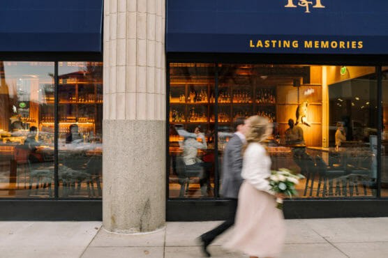 A blurred couple, one holding a bouquet, walks past a bar with large windows and a blue awning that reads LASTING MEMORIES. The bar inside is warmly lit, with patrons seated at the counter. by Justin Salem Meyer