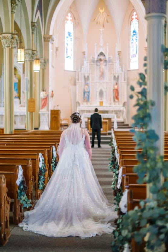 A bride in a white gown walks down the aisle of a church decorated with greenery, approaching a groom standing near the altar. The church has tall arches, stained glass windows, and ornate altar details. by Justin Salem Meyer
