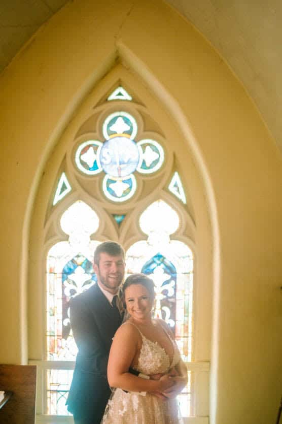 A bride and groom smiling and embracing in front of a tall, ornate stained glass window inside a church. Warm light filters through the colorful glass, highlighting the couples joyful expressions. by Justin Salem Meyer
