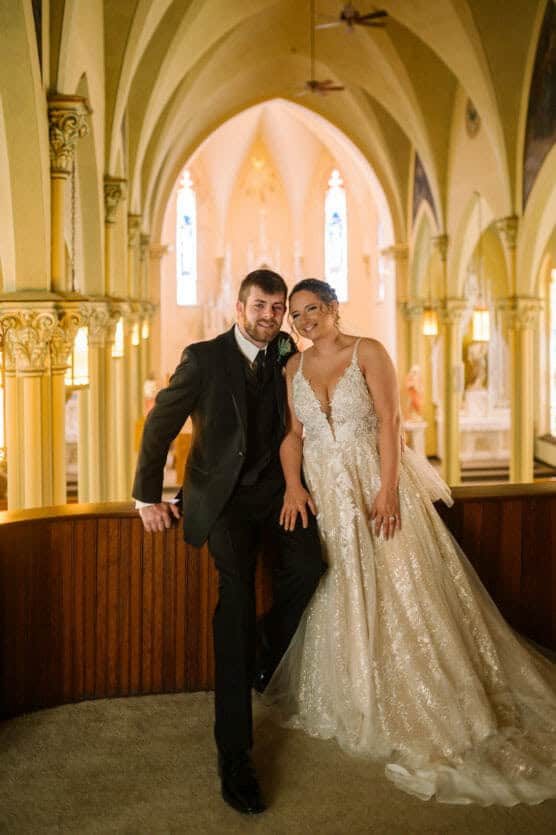 A bride in a glittering white gown and a groom in a black suit pose together inside a beautifully lit church with tall arches and stained-glass windows. by Justin Salem Meyer