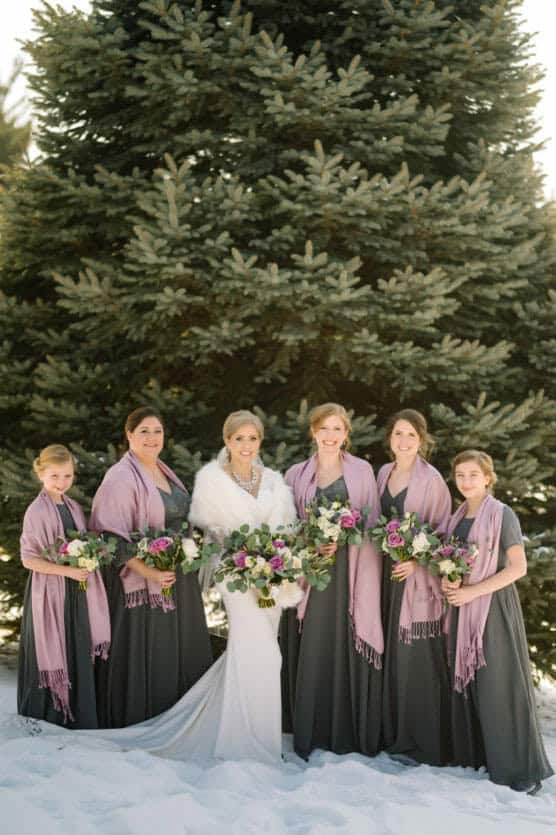 A bride in a white dress and fur wrap stands with five bridesmaids in dark dresses and pink shawls, all holding bouquets, posing in the snow in front of a large evergreen tree. by Justin Salem Meyer