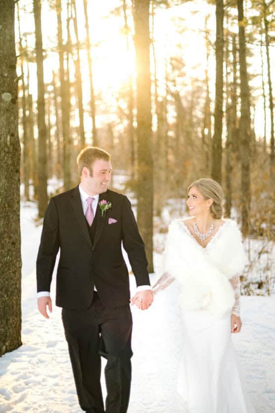 A bride and groom hold hands and smile at each other while walking through a snowy forest with sunlight streaming through the trees. The bride wears a white dress and fur stole; the groom wears a black suit and lavender tie. by Justin Salem Meyer