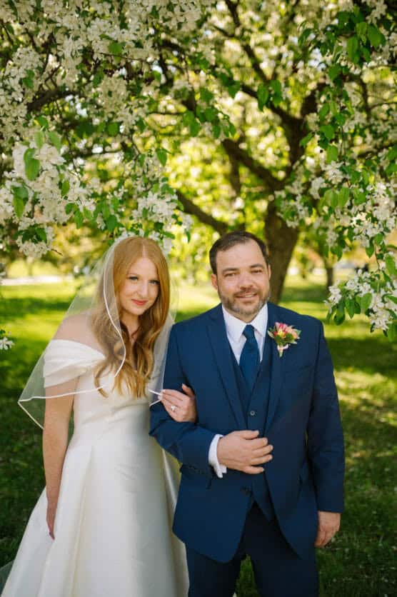 A bride in an off-shoulder white dress and veil stands arm-in-arm with a groom in a blue suit, posing under a blossoming tree in a sunlit garden. Both are smiling gently at the camera. by Justin Salem Meyer