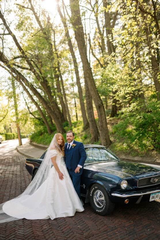 A bride in a white gown and veil stands next to a groom in a blue suit, both smiling beside a vintage dark blue Ford Mustang on a sunlit, tree-lined brick road. by Justin Salem Meyer