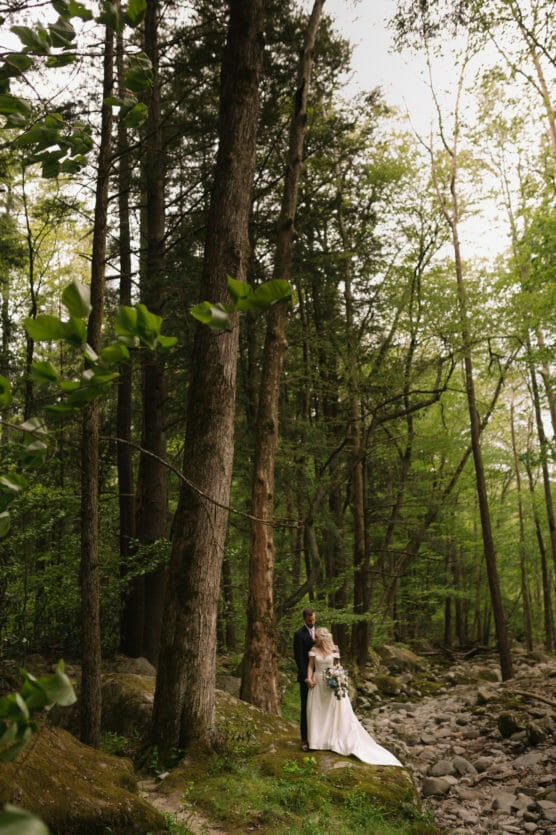 A bride and groom stand together in a lush, green forest beside a rocky stream, surrounded by tall trees and vibrant foliage. The bride wears a white gown and holds a bouquet, while the groom stands behind her. by Justin Salem Meyer