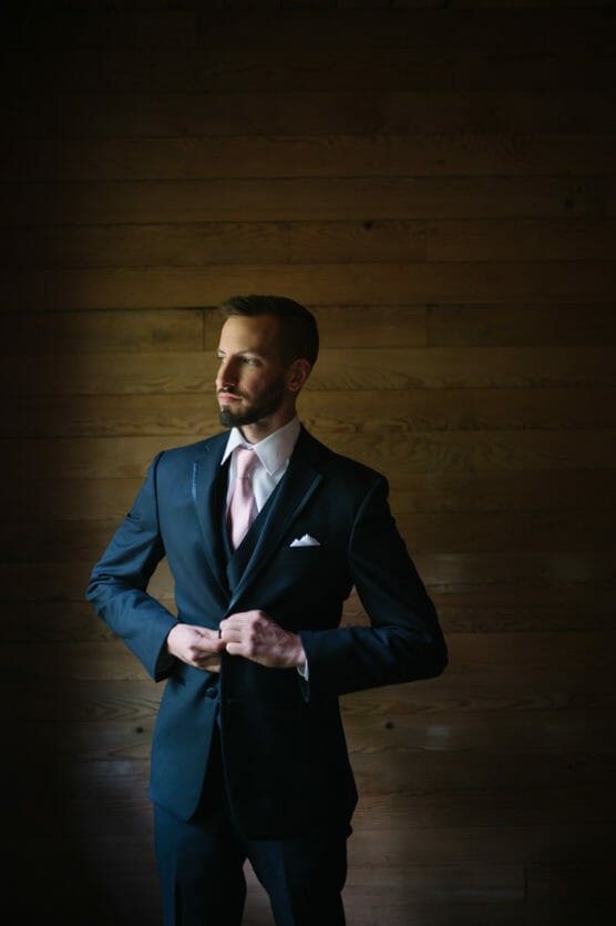 A man in a dark suit with a pale pink tie and pocket square stands in front of a wooden wall, buttoning his jacket and gazing thoughtfully to the left. Soft light highlights one side of his face and body. by Justin Salem Meyer