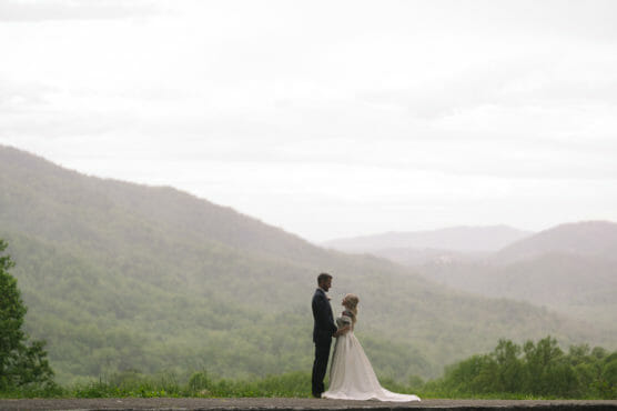 A bride and groom stand facing each other outdoors, surrounded by misty, green mountains under a cloudy sky, with the bride in a white dress and the groom in a dark suit. by Justin Salem Meyer