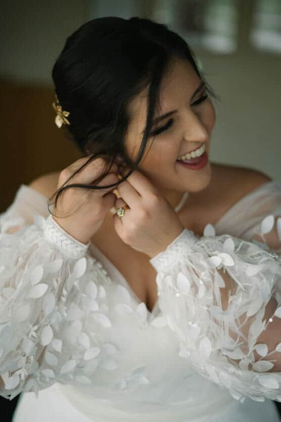 A woman in a white, textured wedding dress smiles joyfully as she touches her earring. Her dark hair is styled up with a decorative clip, and she wears a ring and a pearl necklace. by Justin Salem Meyer
