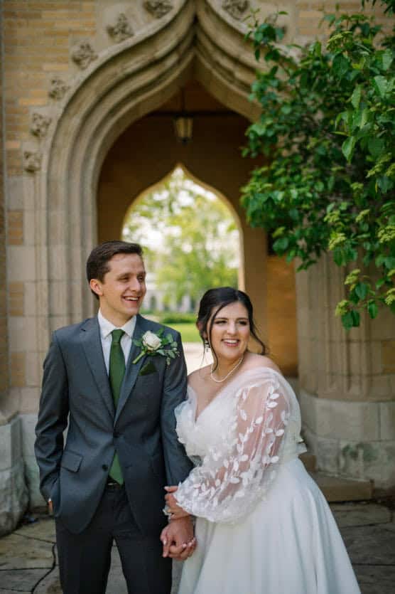 A bride and groom stand smiling under a stone archway, holding hands. The groom wears a gray suit with a green tie and boutonniere; the bride wears a white wedding dress with sheer sleeves. Green foliage is visible nearby. by Justin Salem Meyer