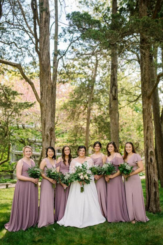 A bride in a white dress stands outdoors with six bridesmaids in matching mauve dresses, all holding bouquets of greenery and flowers, surrounded by tall trees and lush greenery. by Justin Salem Meyer