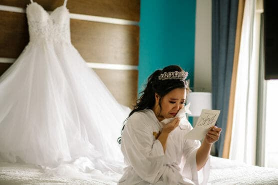 A bride in a white robe and tiara sits on a bed, wiping away a tear while reading a letter. Her white wedding dress hangs in the background. Light comes in from a window beside her. by Justin Salem Meyer