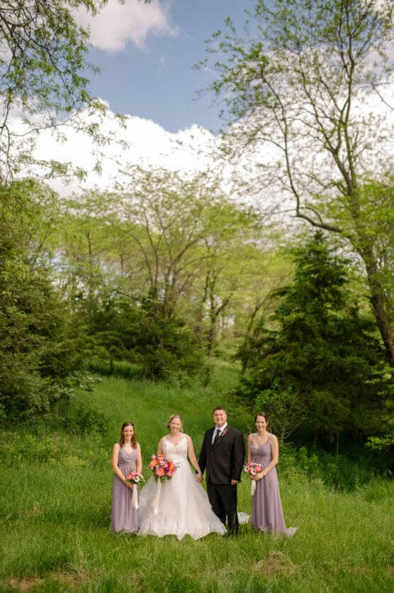 A bride and groom stand hand in hand outdoors, flanked by two bridesmaids in lavender dresses, all holding bouquets, surrounded by lush green trees and grass under a partly cloudy sky. by Justin Salem Meyer