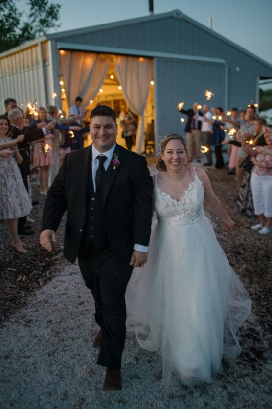 A bride and groom walk hand in hand, smiling, outside a barn venue at dusk. Guests line the path on both sides, holding sparklers in celebration. The scene is festive and joyful. by Justin Salem Meyer