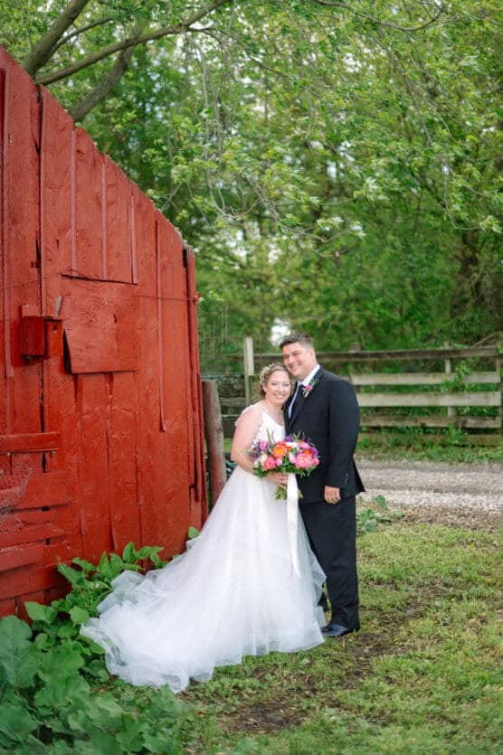 A bride and groom stand smiling together outdoors by a large red wooden barn, surrounded by greenery. The bride holds a colorful bouquet and wears a white gown, while the groom is dressed in a dark suit and tie. by Justin Salem Meyer