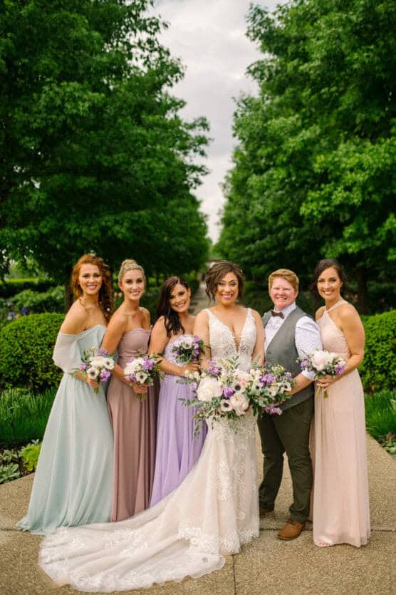 A group of six people dressed in formal attire pose outdoors on a path, surrounded by greenery. The person in a white wedding dress stands in the center, holding a bouquet, with five attendants in pastel dresses and one in a vest and bow tie. by Justin Salem Meyer