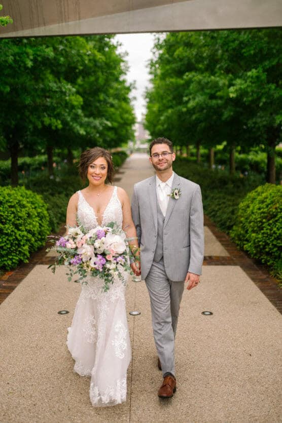 A bride in a white lace gown holding a bouquet walks hand-in-hand with a groom in a light gray suit along a garden pathway lined with green trees, both smiling at the camera. by Justin Salem Meyer