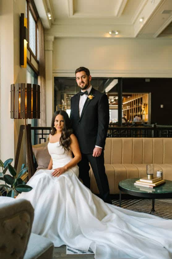 A bride in a white gown sits on a beige sofa while a groom in a black tuxedo stands beside her in a modern, elegant room with large windows and stylish decor. by Justin Salem Meyer