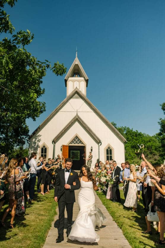 A newlywed couple walks arm in arm down the path from a white chapel, surrounded by guests celebrating and blowing bubbles on a sunny day. by Justin Salem Meyer