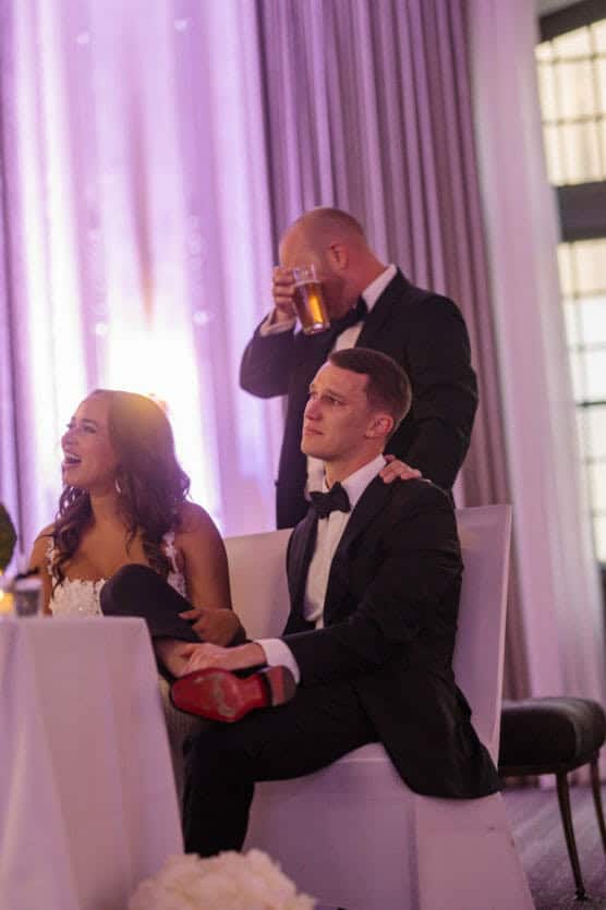 A bride and groom sit together at a reception, the bride smiling with her legs draped over the grooms lap. The groom looks thoughtful. A man in a suit stands behind them, drinking from a glass. The room is softly lit with purple tones. by Justin Salem Meyer