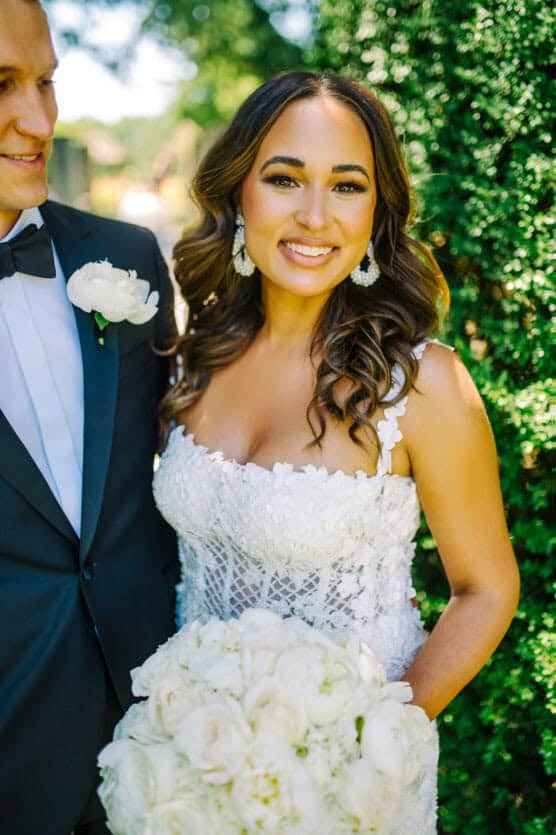 A smiling bride in a white lace wedding dress holds a bouquet of white flowers, standing outdoors next to a man in a suit with a white rose boutonniere. Lush greenery is in the background. by Justin Salem Meyer