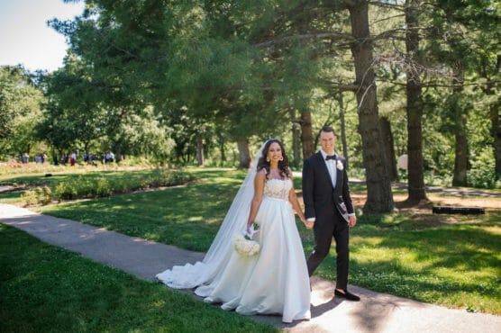 A bride in a white gown and a groom in a black tuxedo walk hand-in-hand on a sunny path through a green park, surrounded by tall trees and lush grass. The bride holds a bouquet of white flowers and smiles brightly. by Justin Salem Meyer