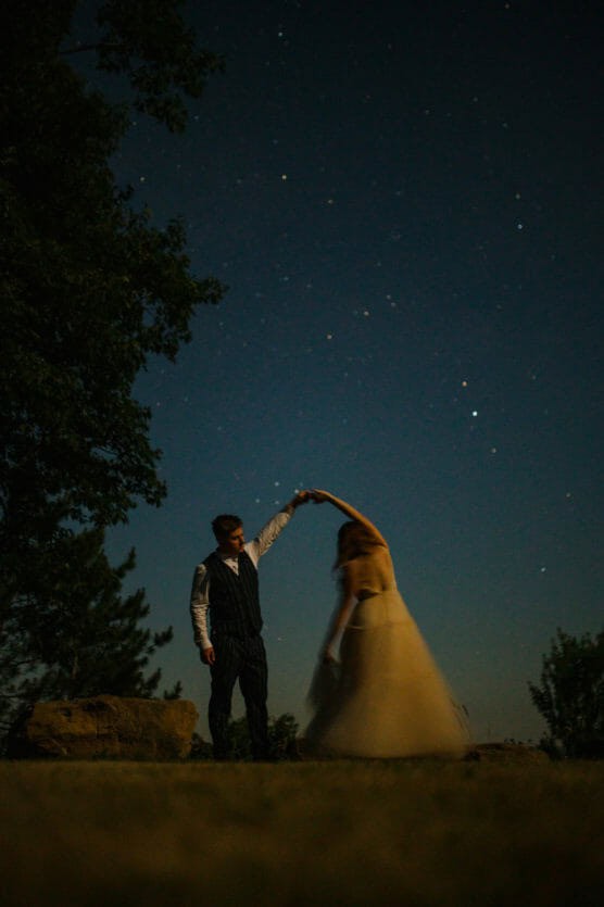 A couple dances outdoors at night under a starry sky. The woman’s dress flows as she spins, while the man holds her hand. Trees and rocks are visible in the dim, serene setting. by Justin Salem Meyer
