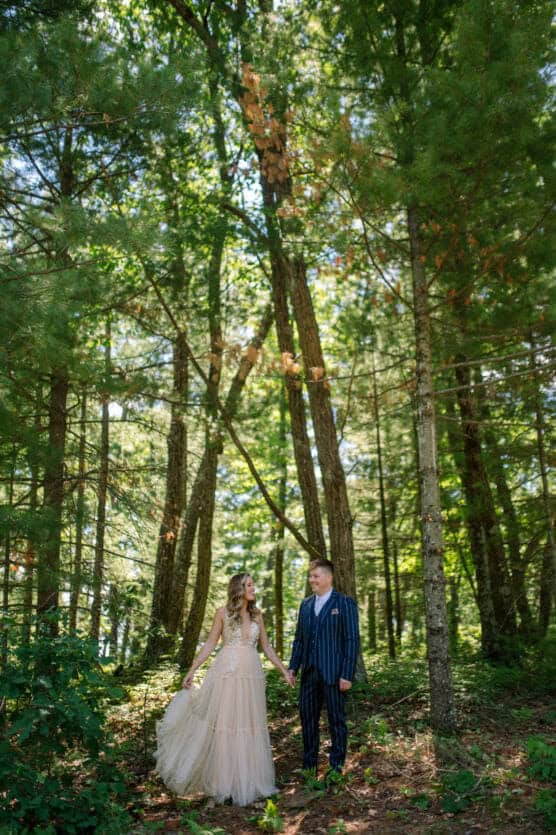 A couple stands hand in hand in a sunlit forest. The woman wears a long, flowing beige dress, while the man is dressed in a navy striped suit. They smile at each other, surrounded by tall green trees. by Justin Salem Meyer