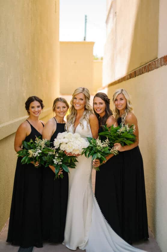 A bride in a white dress stands smiling with four bridesmaids in black dresses, all holding green and white bouquets, in a narrow sunlit outdoor corridor with beige walls. by Justin Salem Meyer