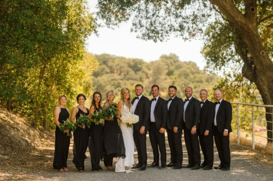 A wedding party poses outdoors on a sunny day, with bridesmaids in black dresses holding bouquets and groomsmen in black suits. The bride in white and groom in black stand at the center, surrounded by trees. by Justin Salem Meyer