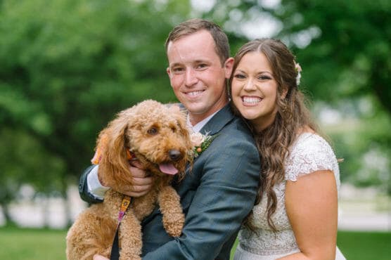 A smiling bride and groom pose outdoors, holding a happy brown curly-haired dog. The couple looks joyful, and the background is filled with green trees. by Justin Salem Meyer